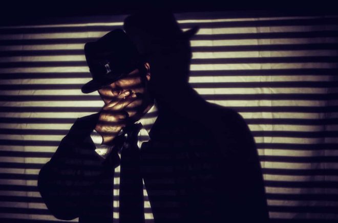 Film Noir Man Smoking Cigarette Shadows. Man in white shirt, black tie and hat standing smoking a cigarette in shadows of blinds, in film noir style.